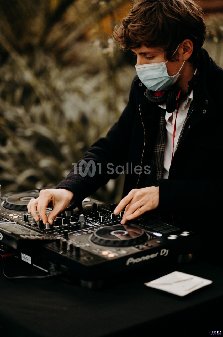 Un DJ portant un masque ajuste les commandes d'une table de mixage dans un environnement extérieur.
