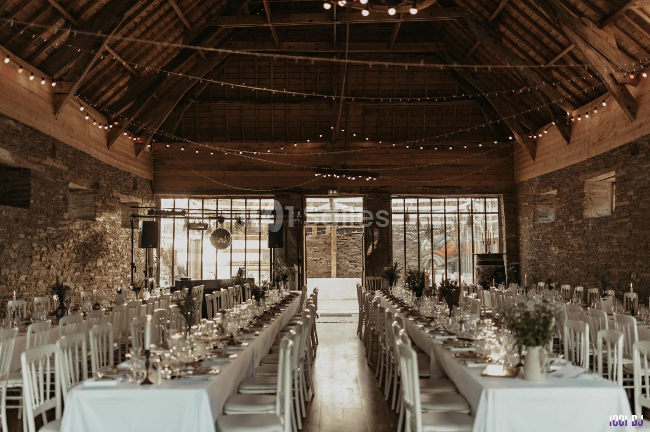 Grande salle en pierre avec tables dressées pour un banquet, éclairée par des guirlandes lumineuses suspendues.