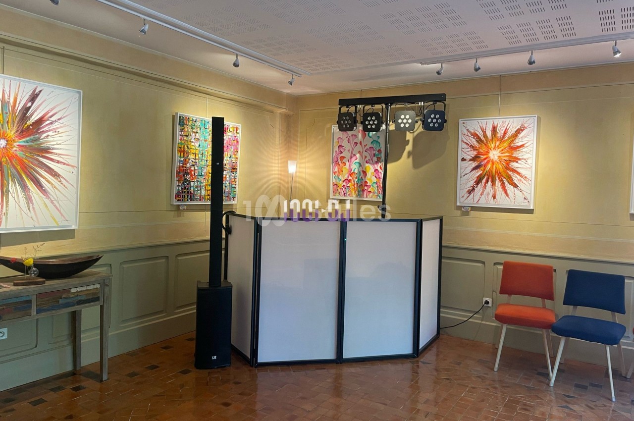 Salle d'exposition avec des peintures colorées accrochées aux murs, un pupitre et des chaises disposées autour.