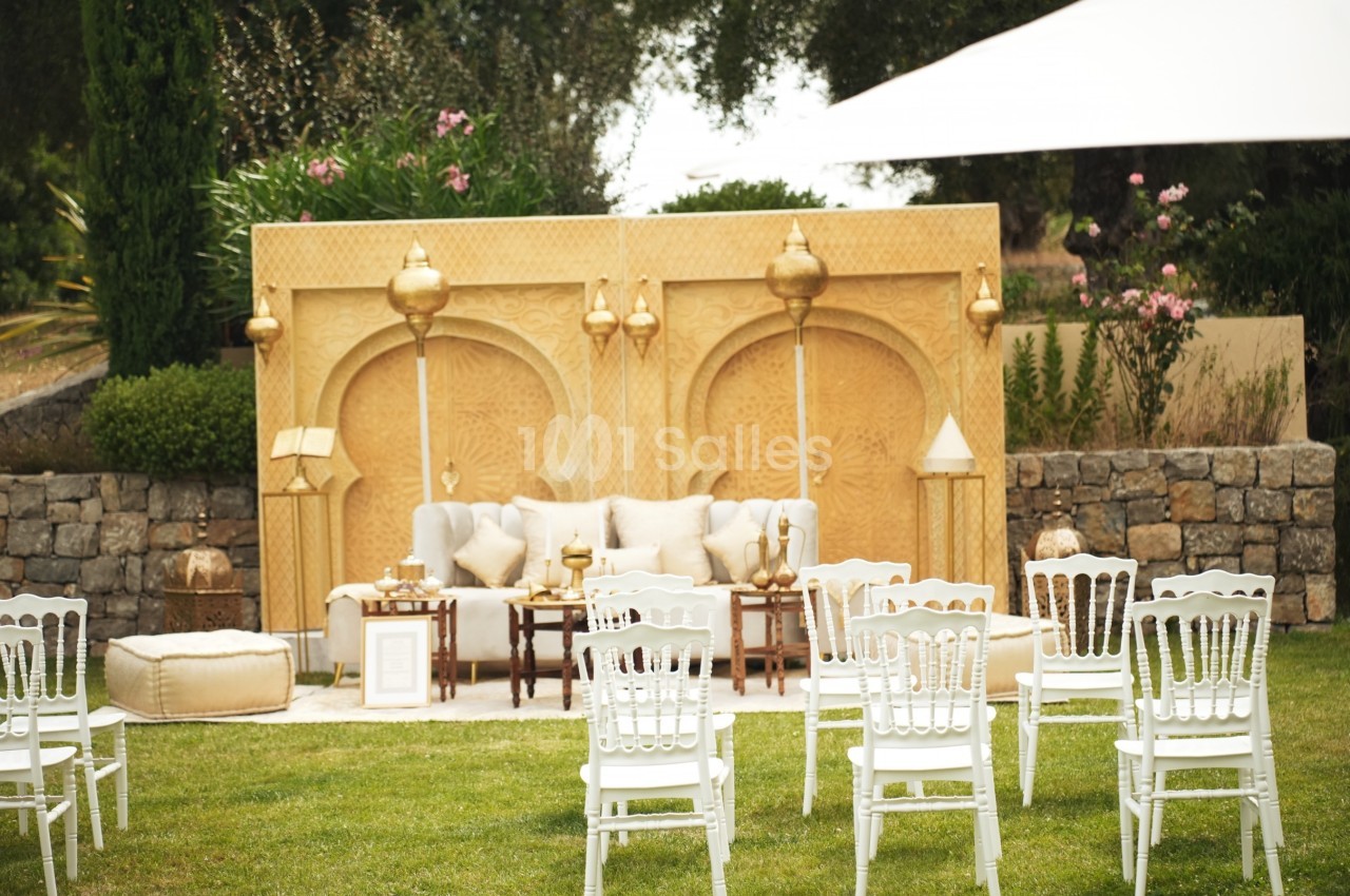 Décoration extérieure avec canapé, coussins et lanternes dorées devant un panneau sculpté, entourée de chaises blanches.