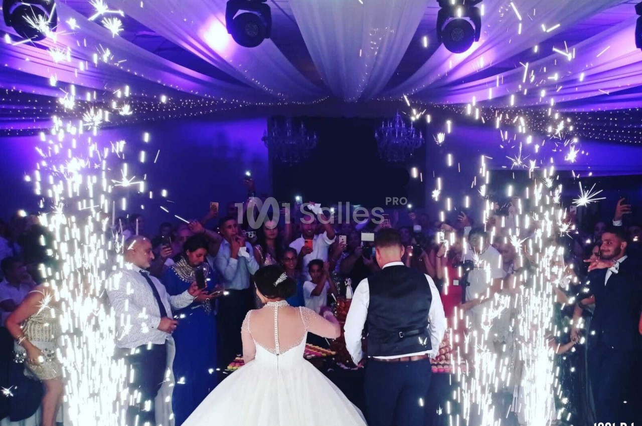 Un couple de mariés coupe leur gâteau sous des étincelles, entouré d'invités dans une salle décorée de lumières violettes.