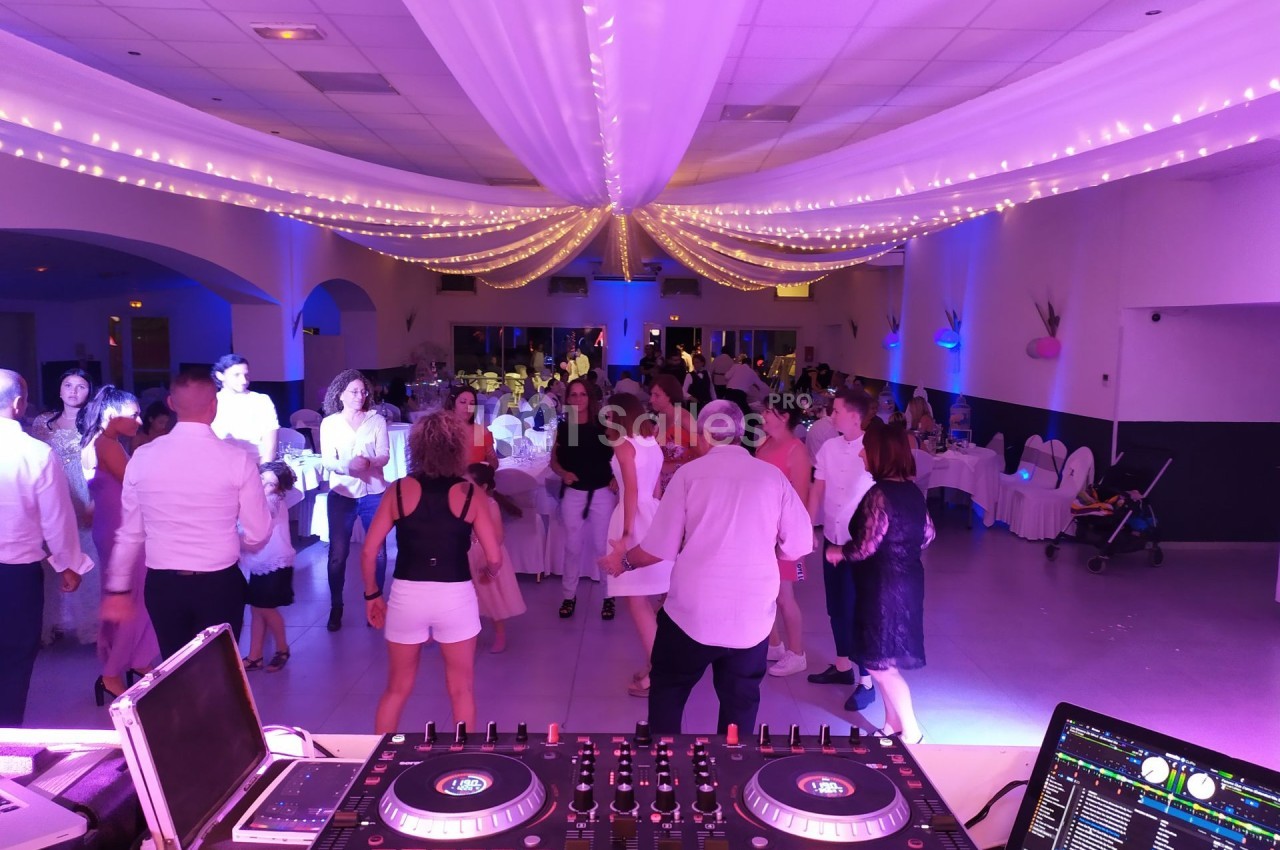 Des personnes dansent dans une salle décorée de guirlandes lumineuses, vue depuis une table de mixage de DJ.