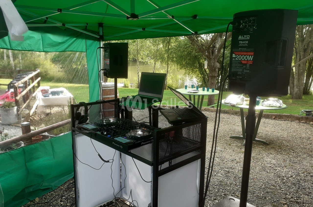 Matériel de DJ installé sous une tente verte, avec platines, ordinateur portable et enceintes, en extérieur près d'un étang.
