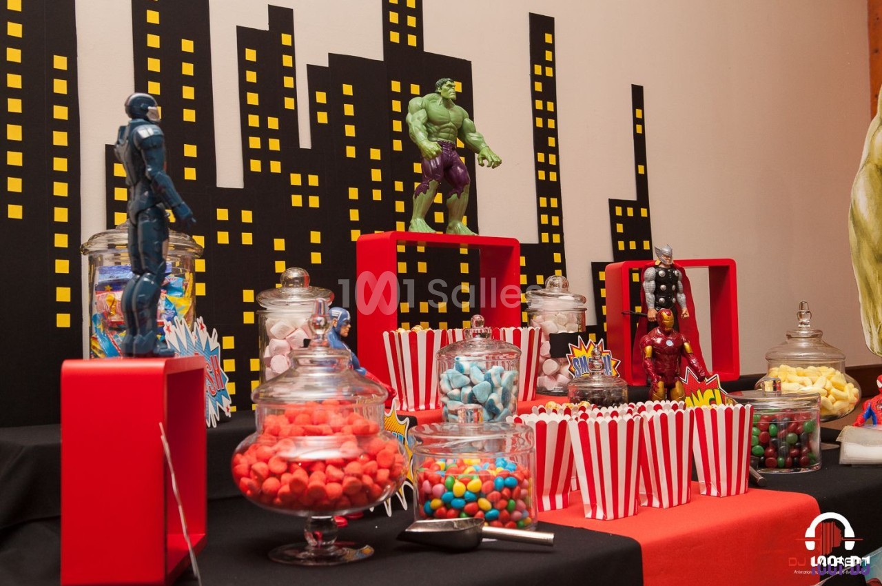 Table décorée sur le thème des super-héros avec bonbons, popcorn et figurines devant un décor de ville stylisé.