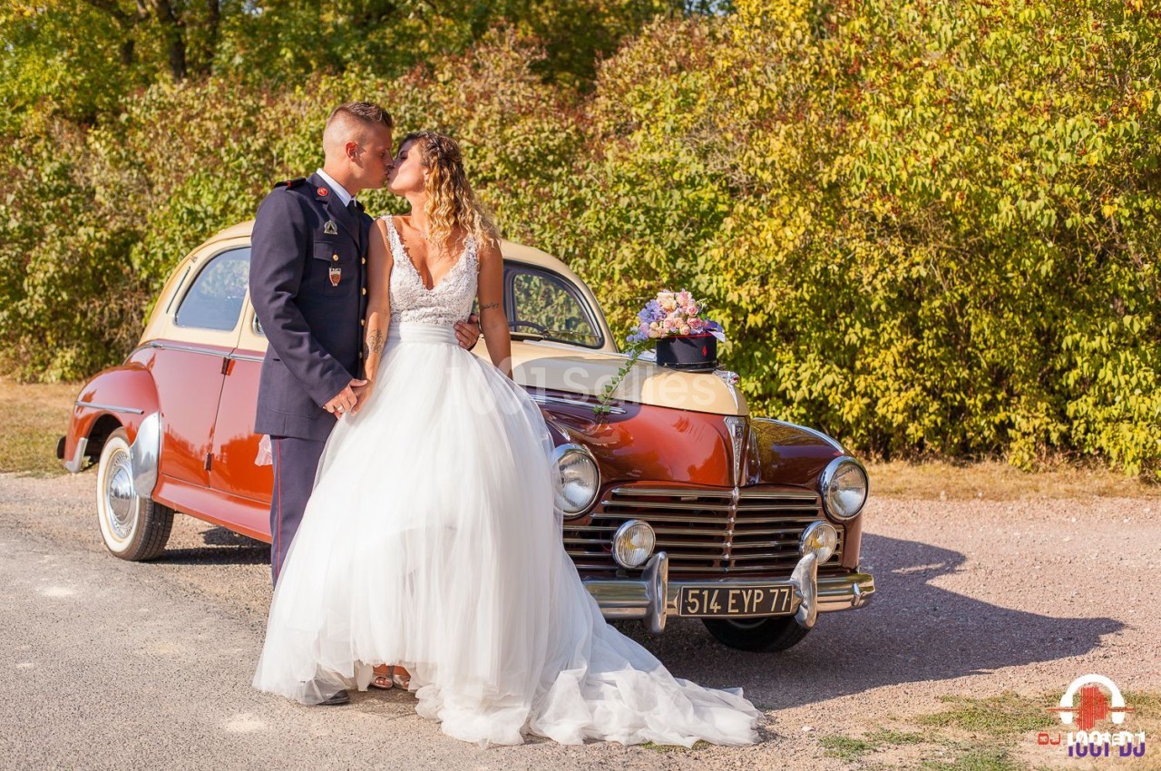 Un couple en tenue de mariage pose devant une voiture ancienne rouge et beige, entouré de végétation.