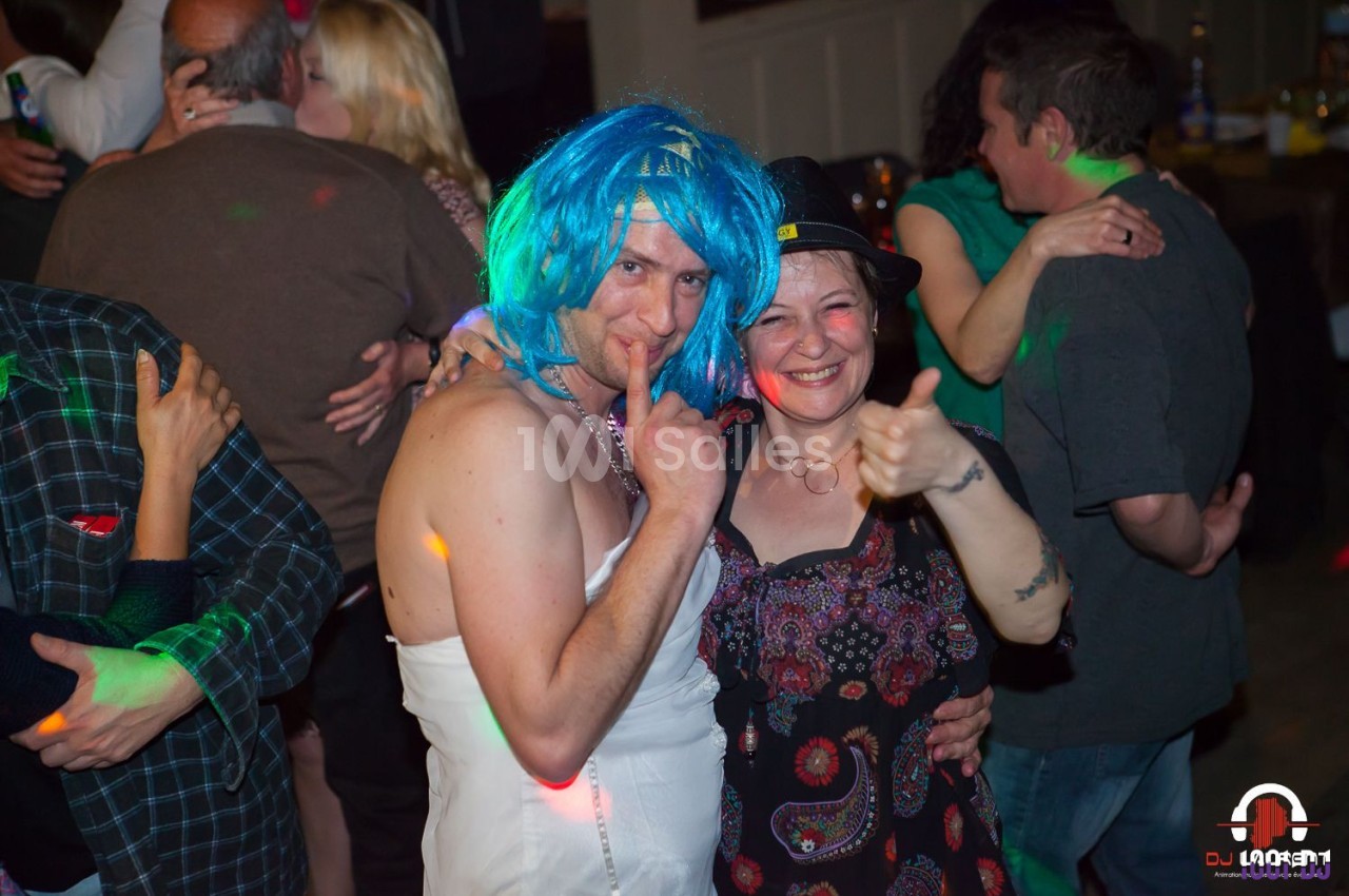 Un homme déguisé en robe blanche et perruque bleue pose avec une femme souriante lors d'une fête animée.
