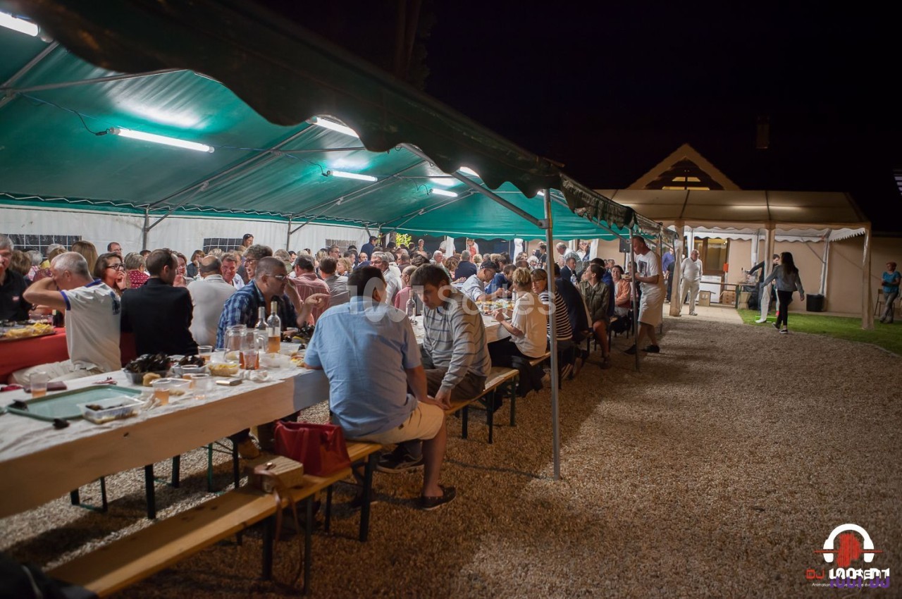 Groupe de personnes dînant sous une tente éclairée en soirée, avec des tables alignées sur un sol en gravier.