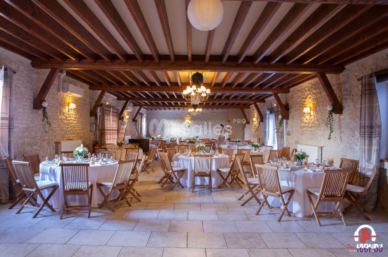 Salle de réception en pierre avec tables rondes dressées, chaises en bois et décoration florale.