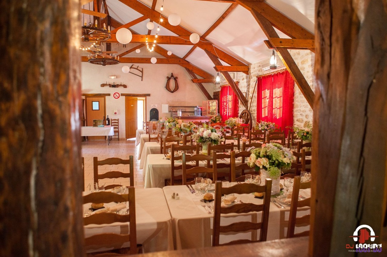 Salle de réception rustique avec tables décorées de fleurs, chaises en bois et poutres apparentes au plafond.