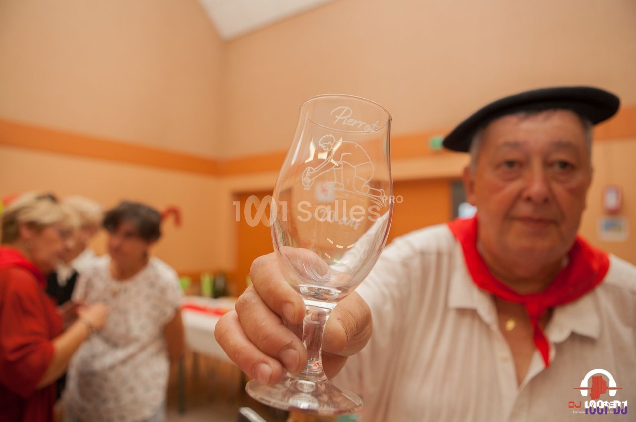 Un homme tenant un verre gravé ’Pierrot 70 ans’ dans une salle avec des personnes en arrière-plan flou.