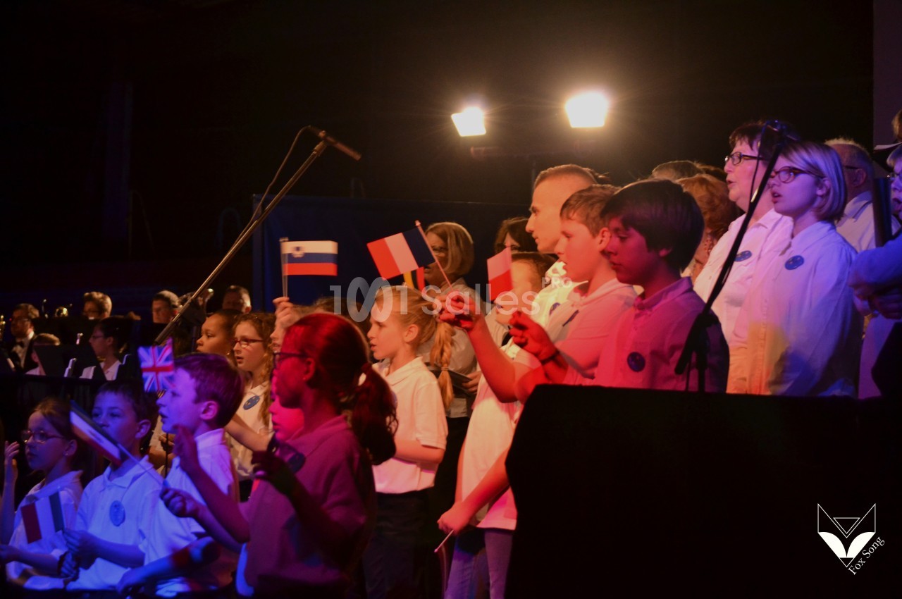 Des enfants et adultes chantent sur scène, tenant des drapeaux de différents pays, éclairés par des projecteurs.