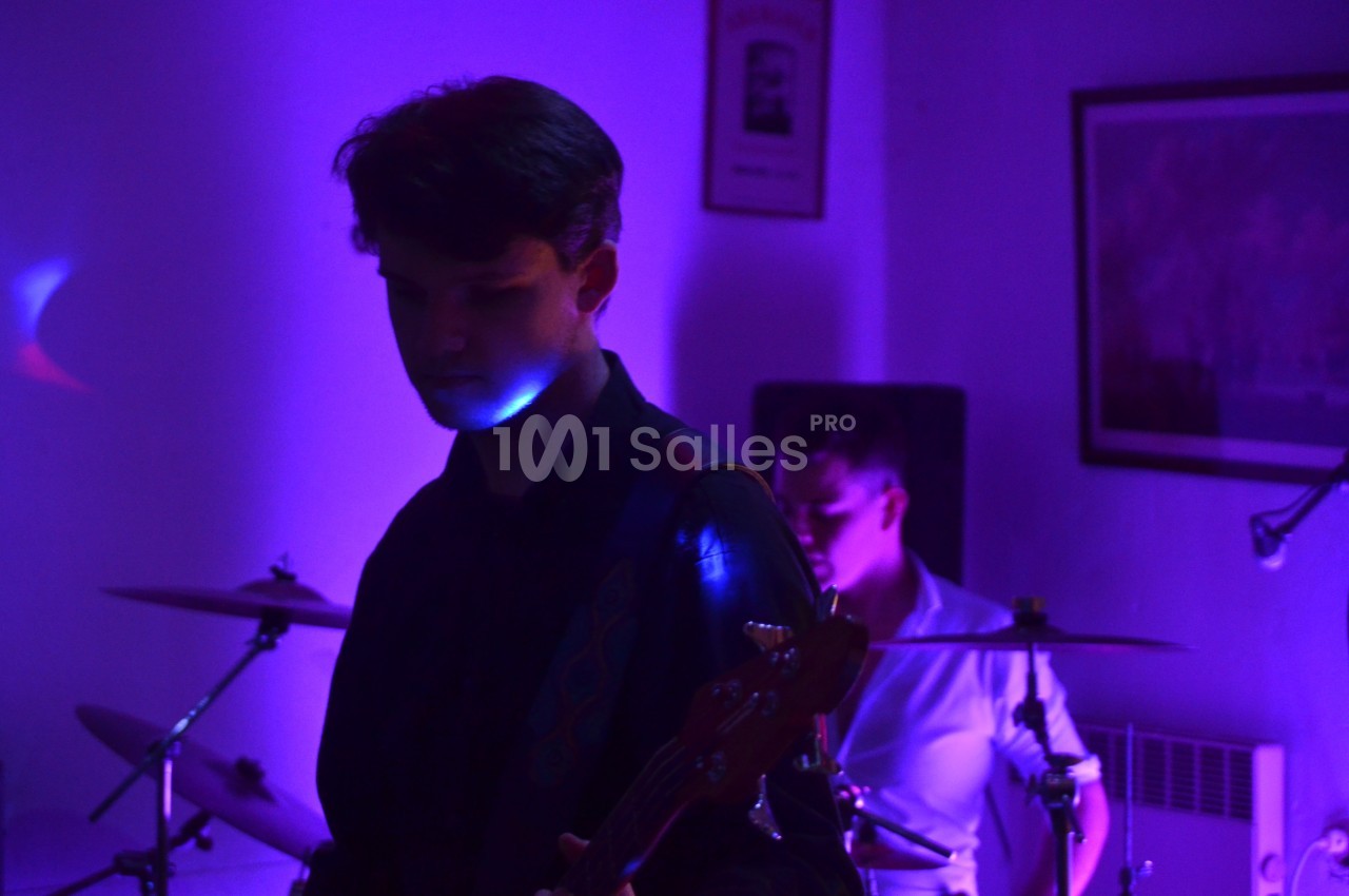Un musicien joue de la guitare basse sous une lumière violette, avec un batteur en arrière-plan.