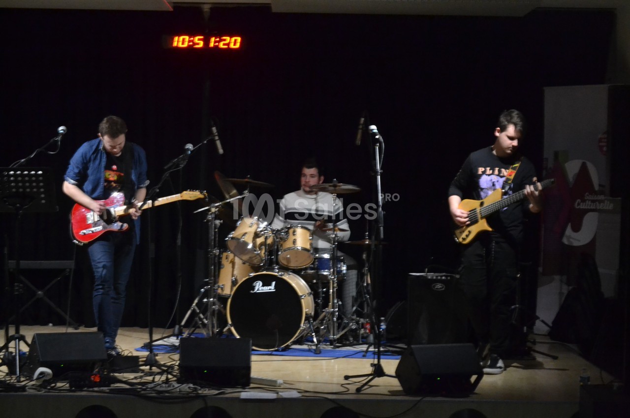 Trois musiciens jouent sur scène avec une guitare électrique, une batterie et une basse, éclairés par une lumière de scène.