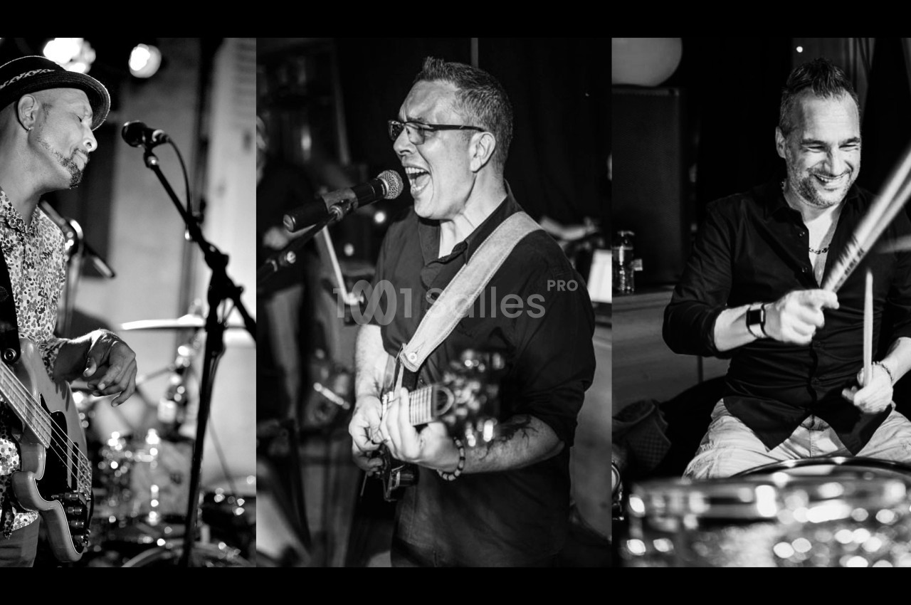 Trois musiciens jouant en concert, avec un bassiste, un chanteur-guitariste et un batteur, en noir et blanc.