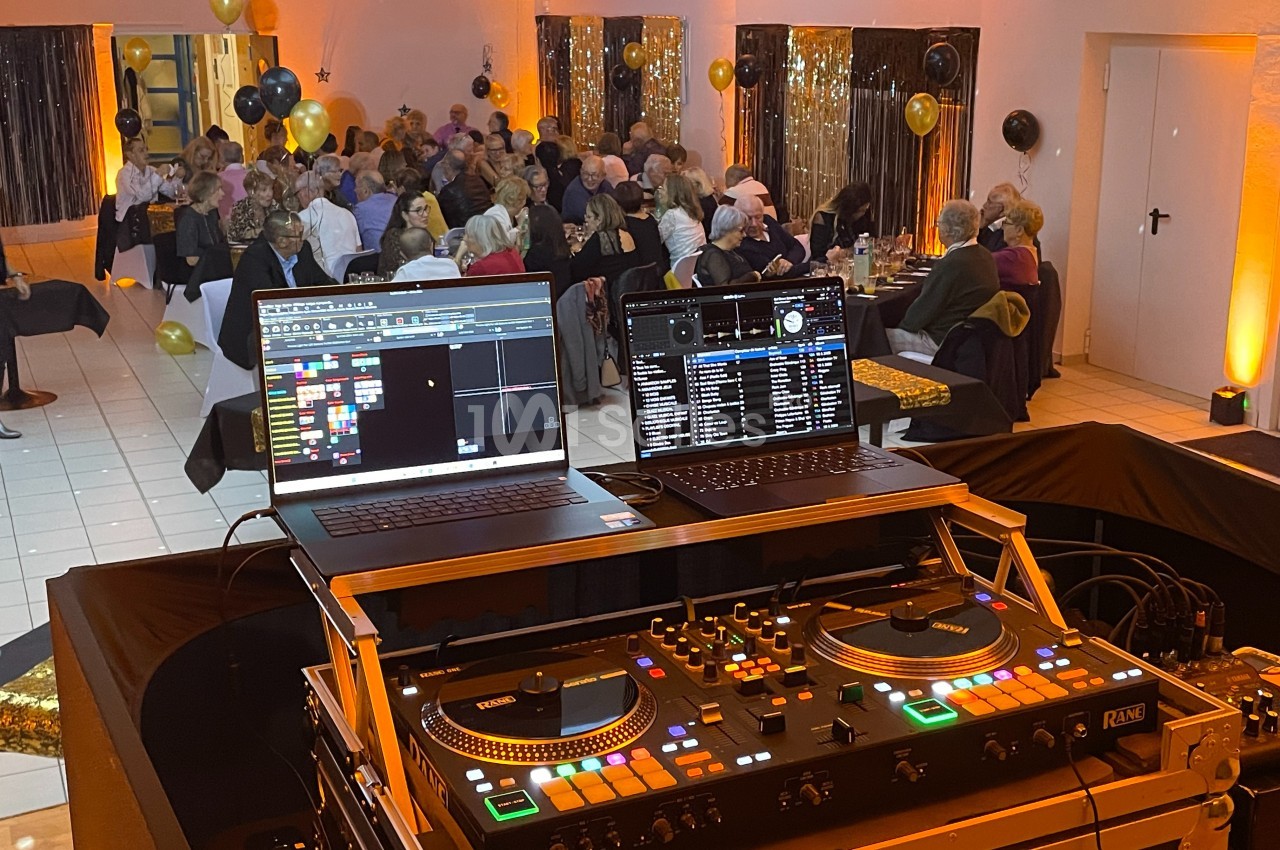 Vue d'une soirée festive avec des invités attablés et une table de mixage DJ au premier plan.