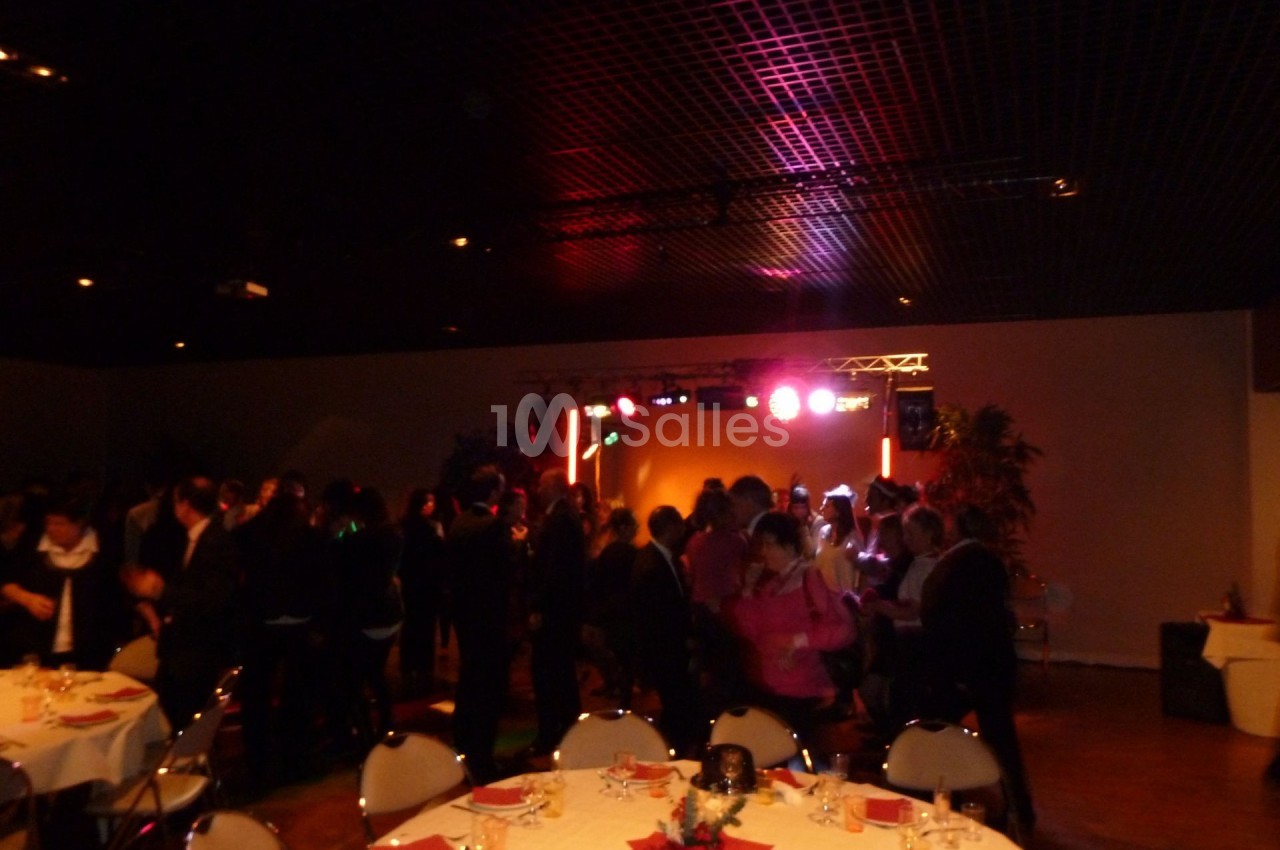 Des personnes dansent dans une salle sombre éclairée par des lumières colorées, avec des tables dressées au premier plan.