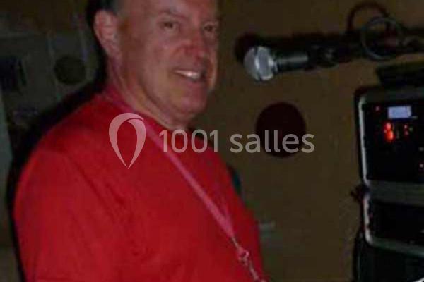 Homme souriant en t-shirt rouge devant un microphone et un équipement audio dans un environnement intérieur sombre.