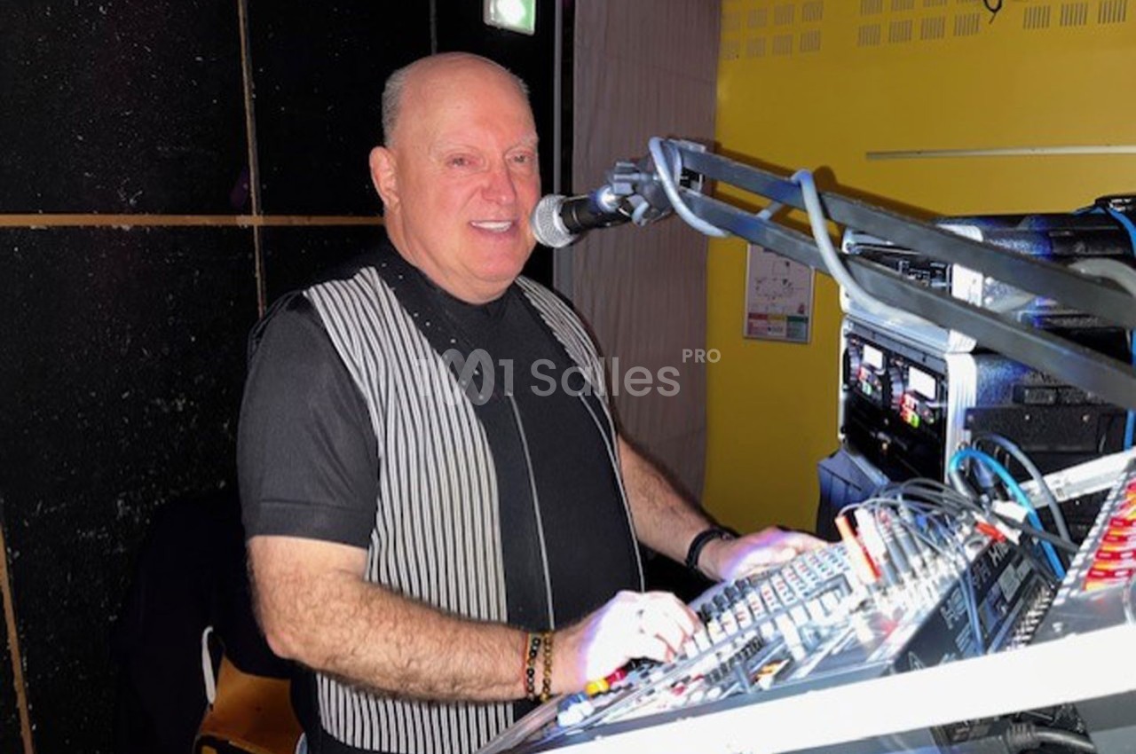 Homme souriant devant une console de mixage, parlant dans un micro, dans un environnement intérieur éclairé.