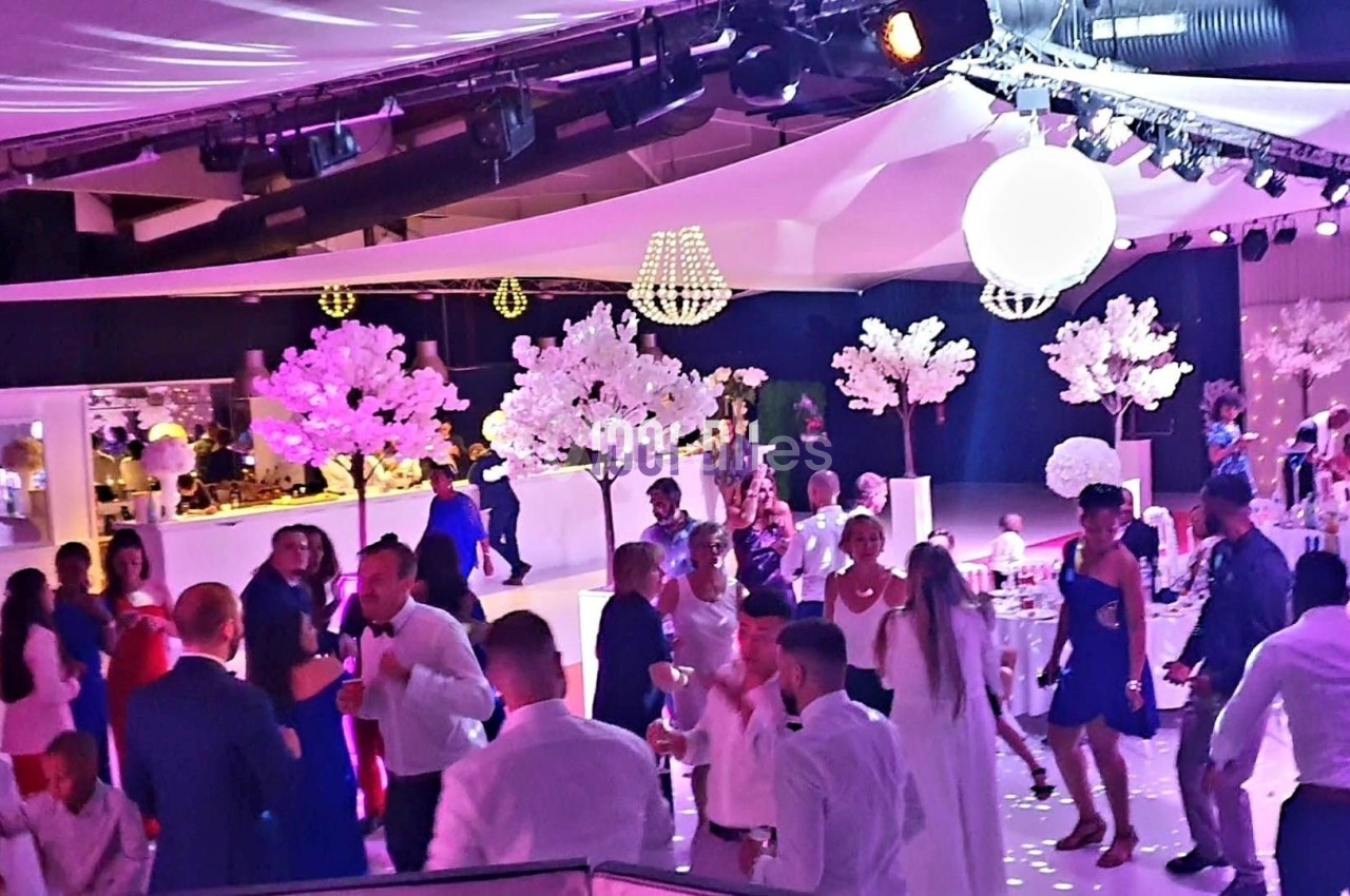 Salle de réception éclairée avec des invités dansant et discutant, décorée de fleurs blanches et lumières suspendues.