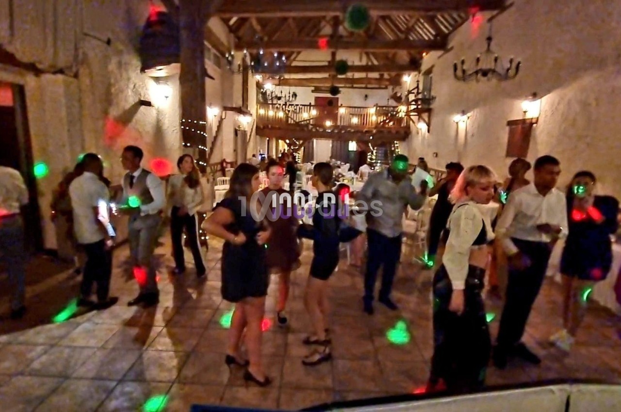 Des personnes dansent dans une salle de réception éclairée par des lumières colorées, avec un décor rustique en arrière-plan.