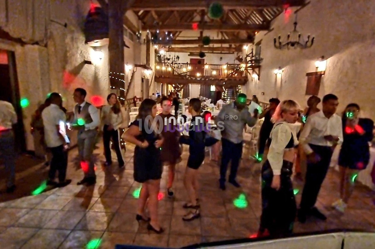 Des personnes dansent dans une salle de réception éclairée par des lumières colorées, avec un décor rustique en arrière-plan.
