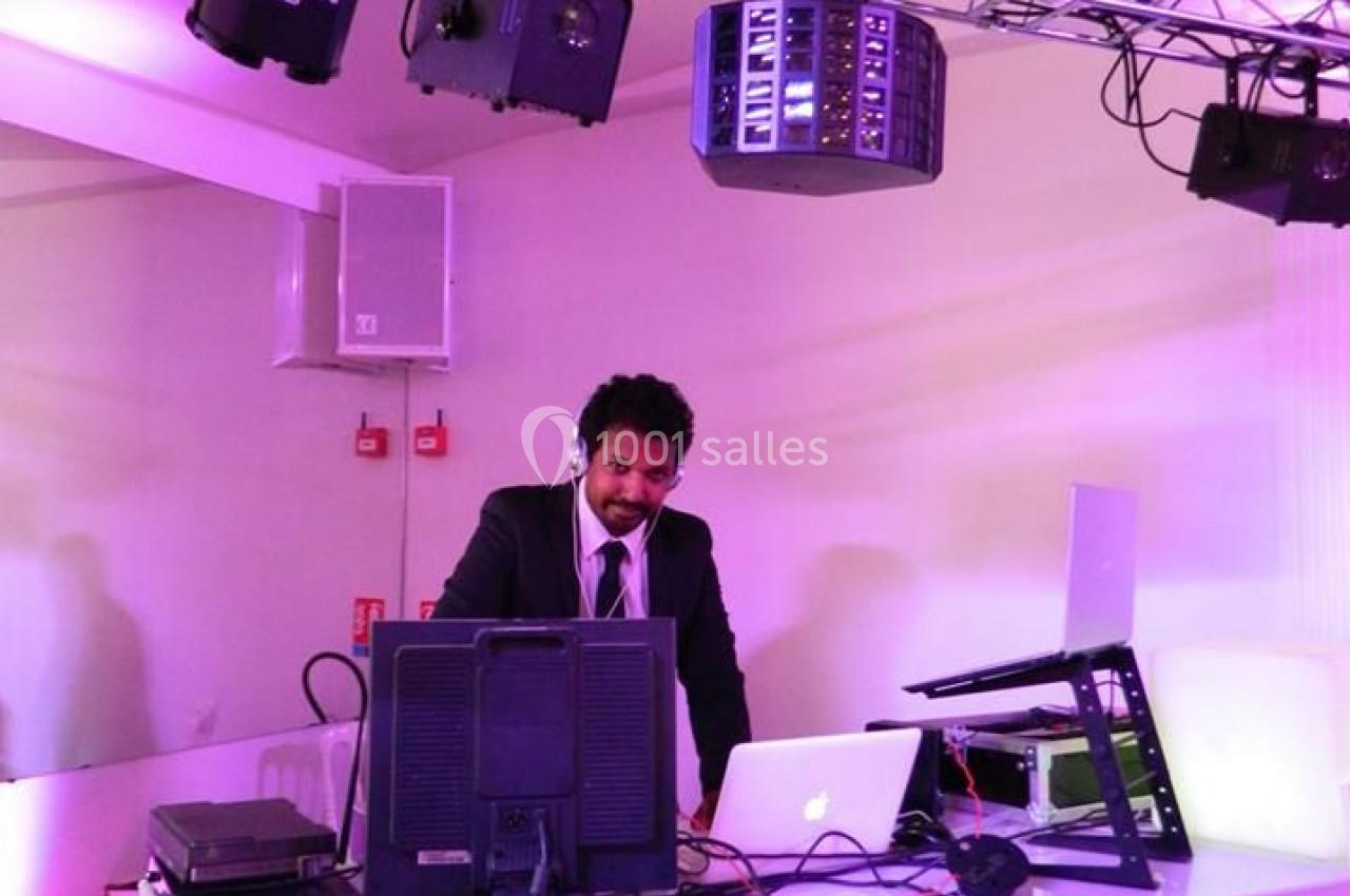 Un DJ en costume mixe de la musique sur une table équipée d'ordinateurs et d'équipements audio sous des lumières violettes.