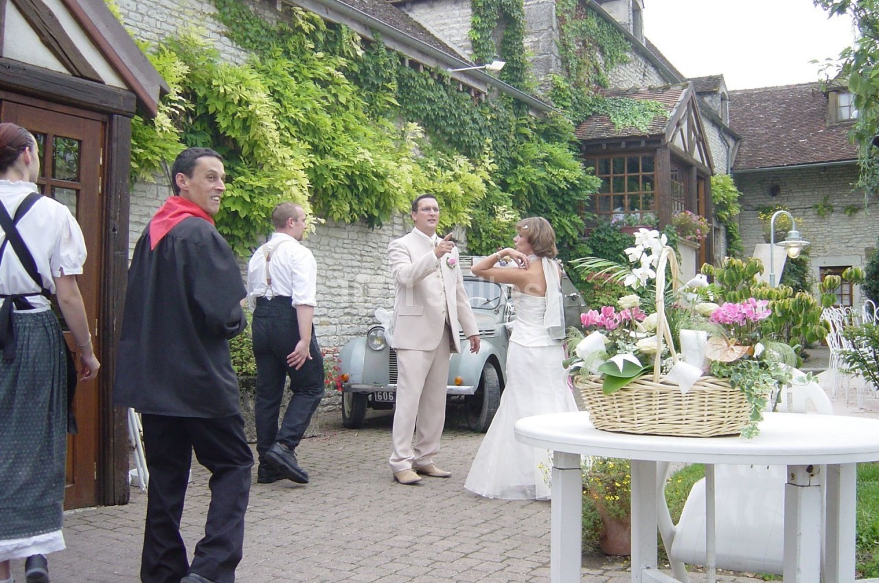 Un couple en tenue de mariage pose devant une voiture ancienne, entouré d'invités dans une cour fleurie.