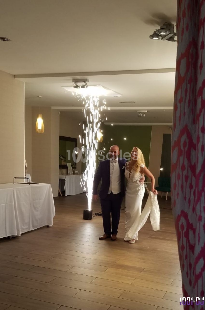Un couple en tenue de mariage entre dans une salle éclairée, accompagné d'effets pyrotechniques en arrière-plan.
