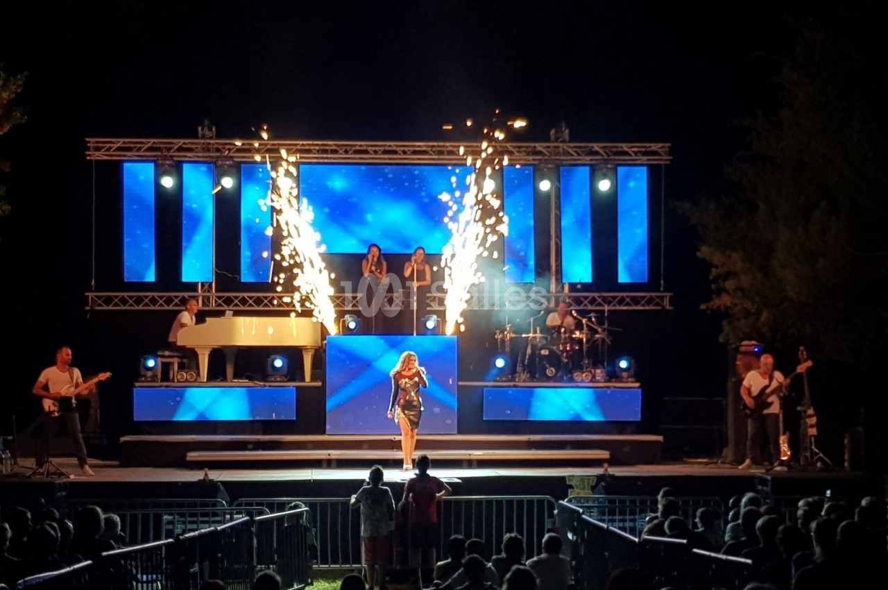 Scène de concert nocturne avec une chanteuse au centre, des musiciens, des écrans lumineux et des effets pyrotechniques.