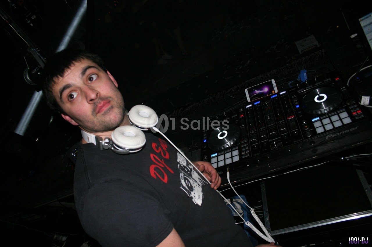 Un DJ portant un casque autour du cou se tient devant une table de mixage dans un environnement sombre.