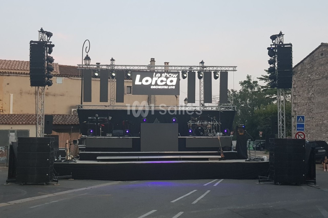 Scène extérieure montée avec éclairages et équipements audio, prête pour un concert en soirée dans un village.
