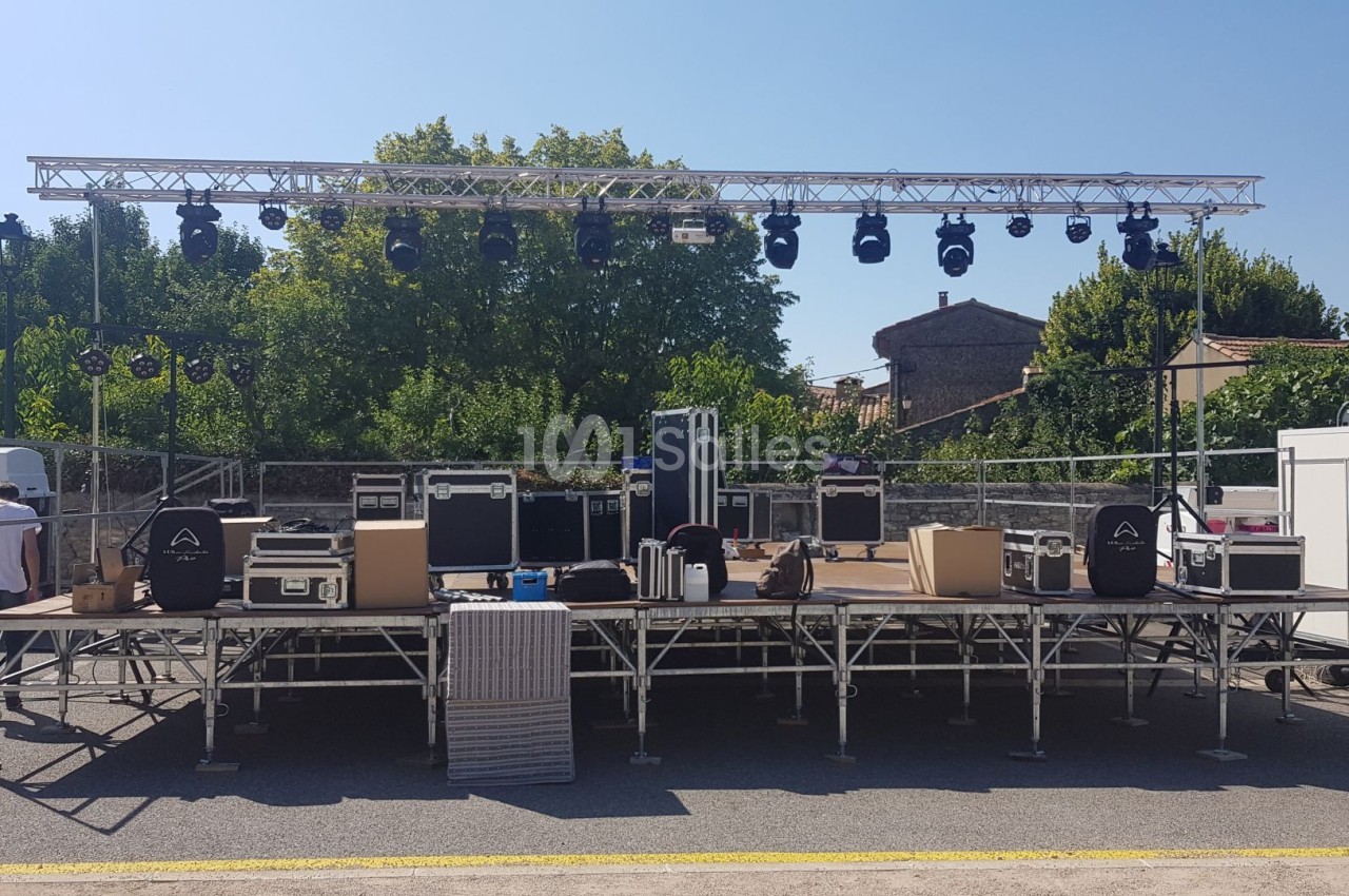 Scène extérieure en montage avec projecteurs suspendus, équipements audio et caisses de transport visibles.