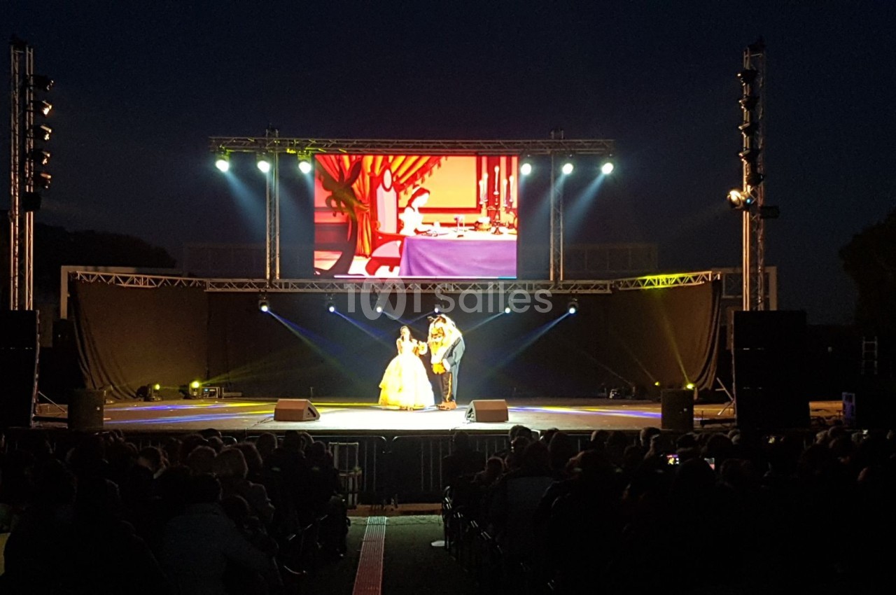 Un spectacle en plein air avec deux artistes sur scène éclairée, devant un écran géant et un public assis.