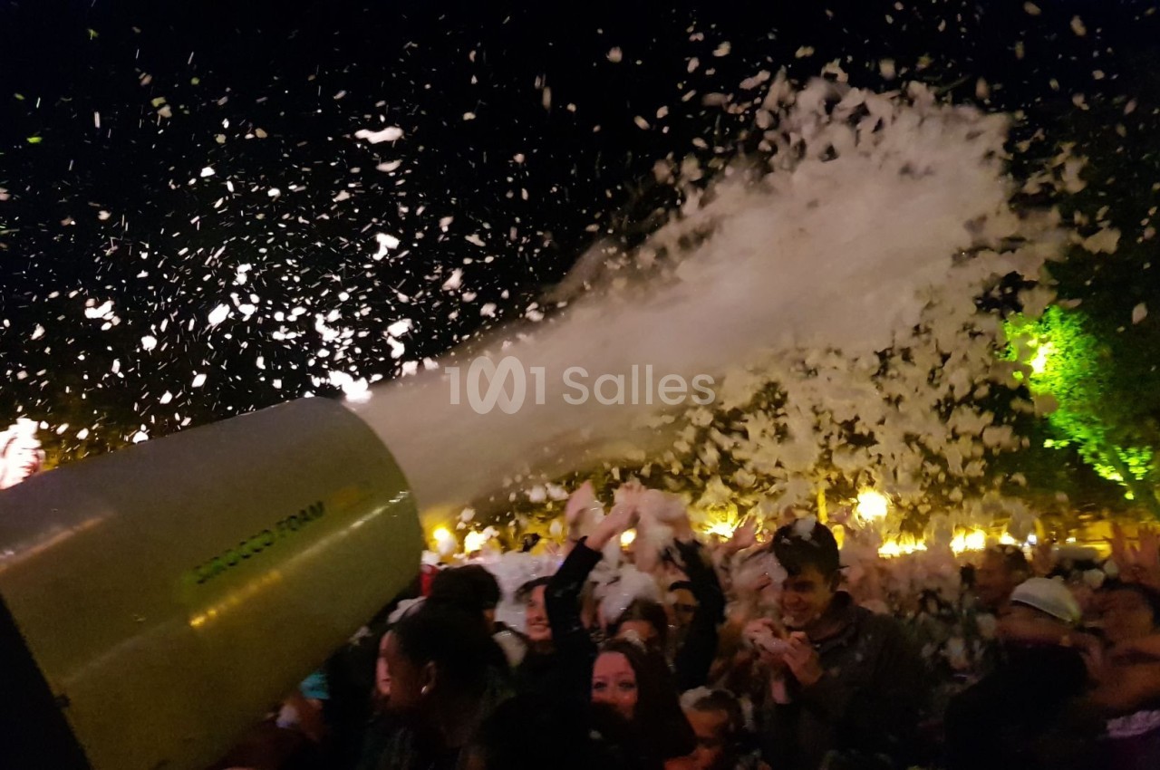 Canon à mousse projetant de la mousse sur une foule en fête, de nuit, avec des lumières en arrière-plan.