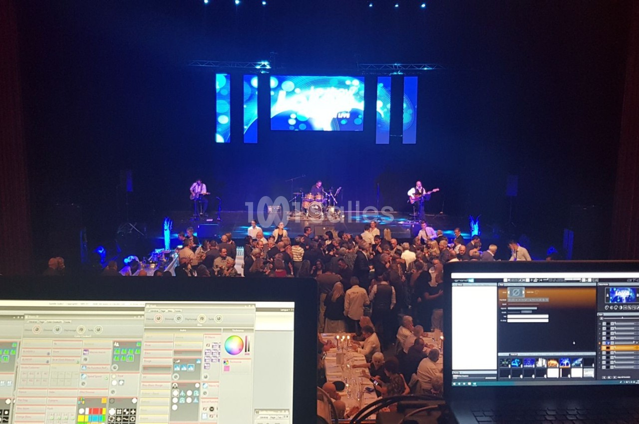 Vue depuis une régie technique sur un concert avec musiciens sur scène, public debout et écrans lumineux en arrière-plan.