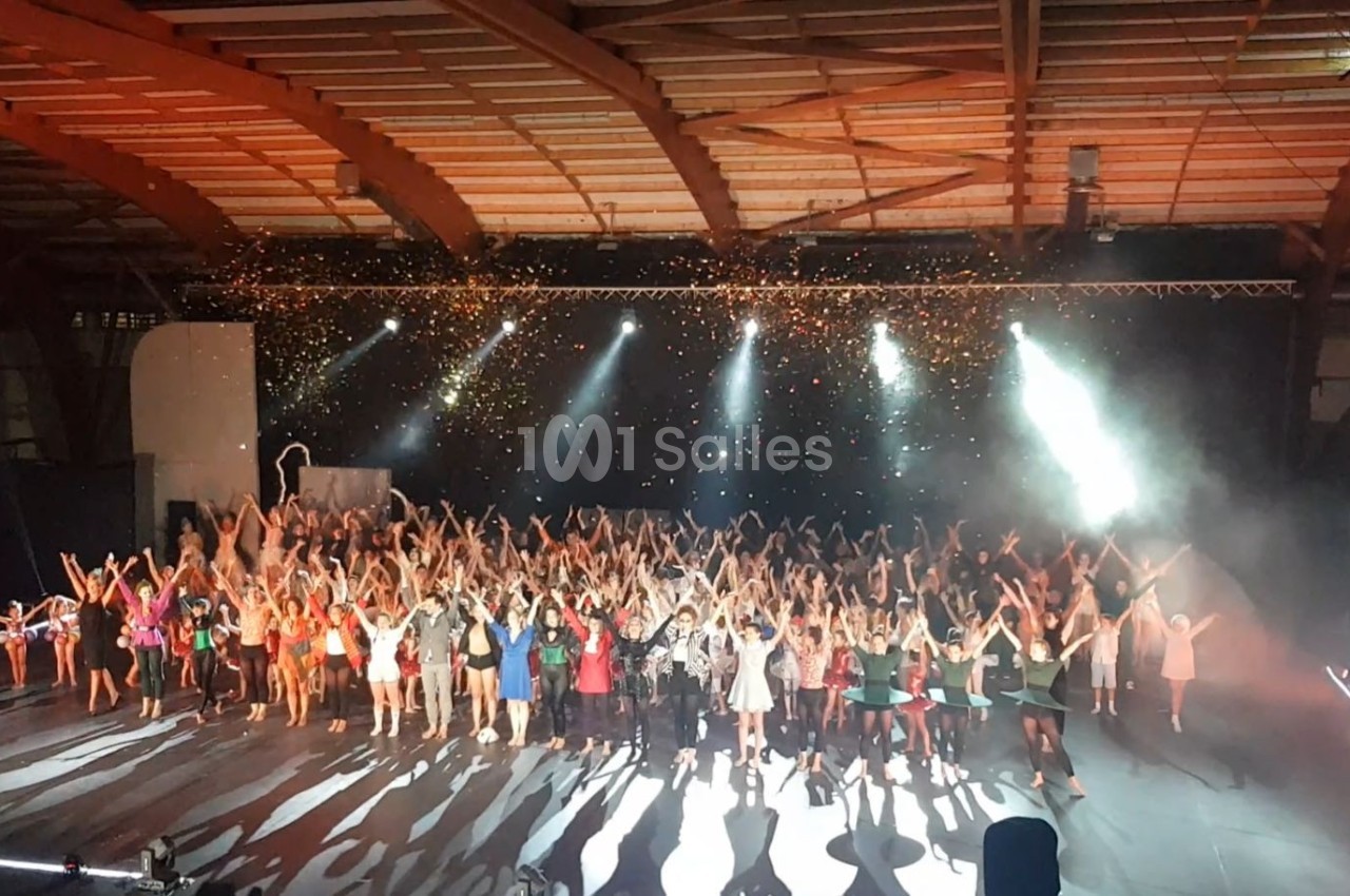 Un grand groupe de personnes sur scène sous des projecteurs et des confettis, dans une salle avec une structure en bois.