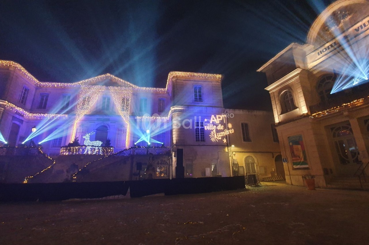 Façade illuminée d'un bâtiment avec décorations de Noël, projecteurs colorés et inscription ’APT ville de lumière’.