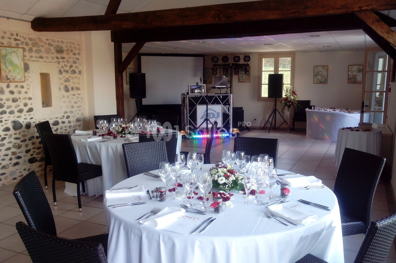 Salle de réception décorée avec des tables rondes dressées, des chaises noires et un espace pour le matériel de DJ.