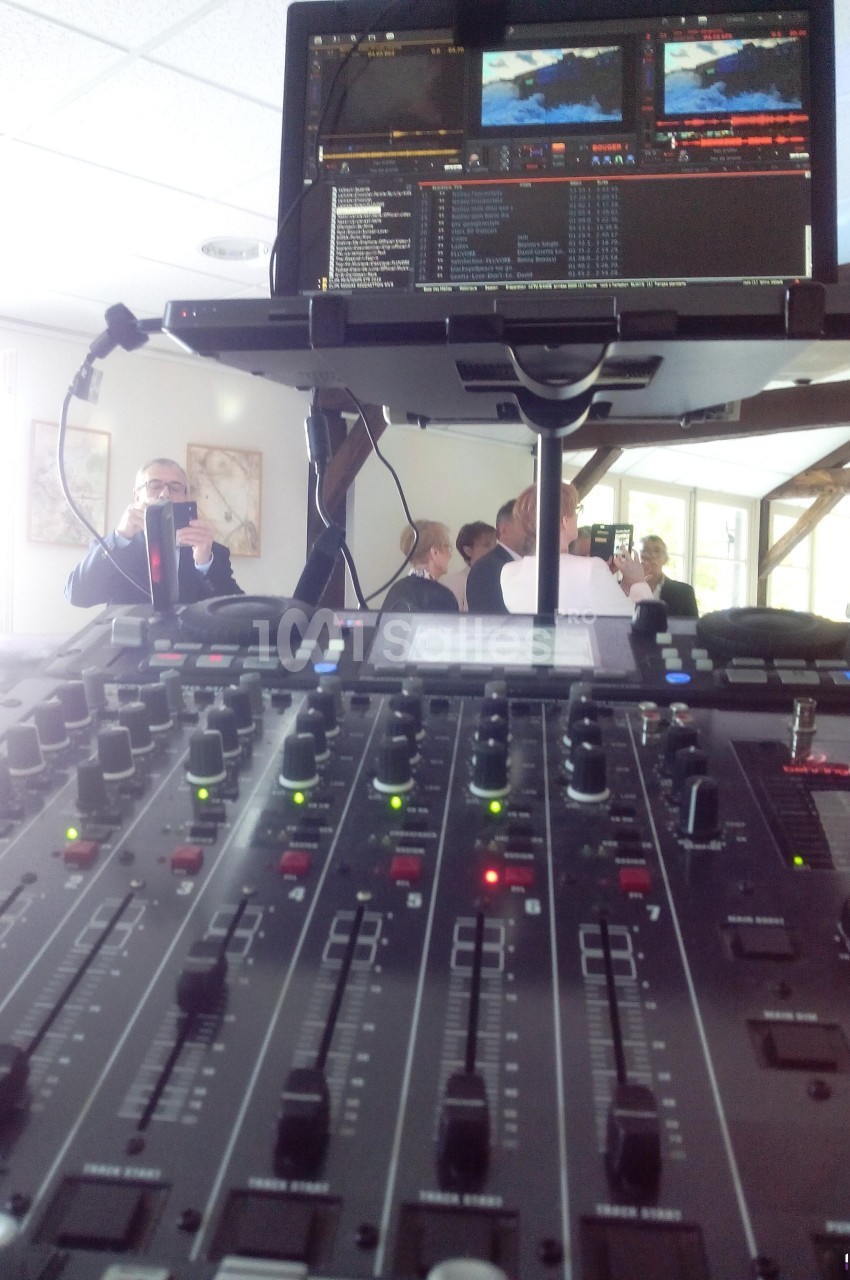 Table de mixage audio en gros plan avec un écran de contrôle vidéo en arrière-plan, dans une salle lumineuse.