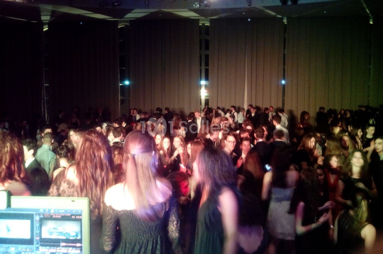 Des personnes dansent dans une salle sombre avec des rideaux fermés, vue depuis une table de mixage.