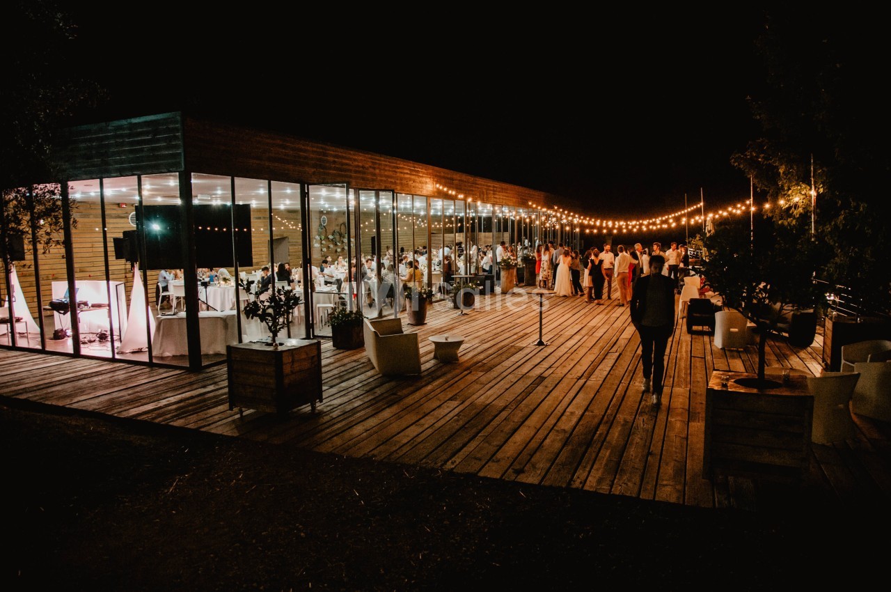 Terrasse en bois éclairée par des guirlandes lumineuses, avec des invités rassemblés près d'un bâtiment vitré.