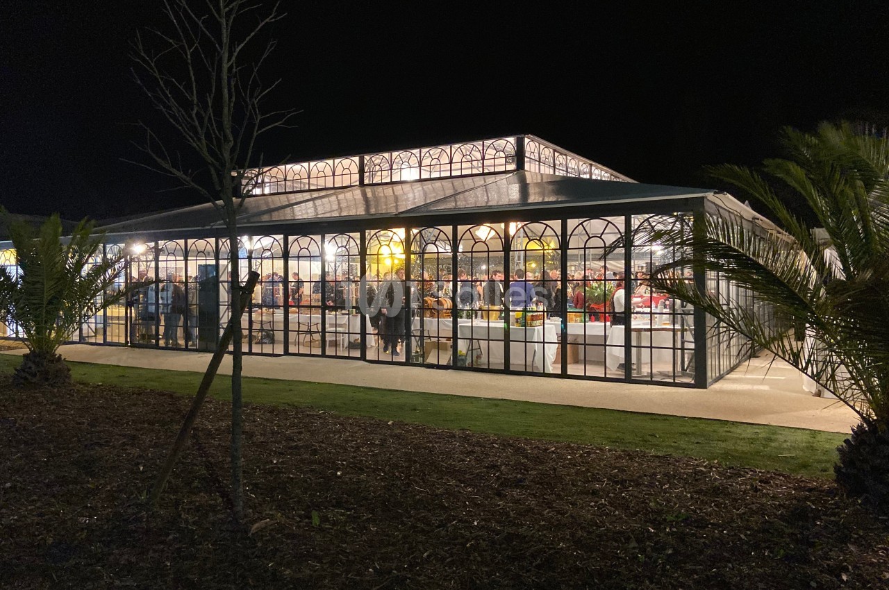 Grande serre illuminée la nuit, avec des personnes visibles à l'intérieur et des plantes autour.