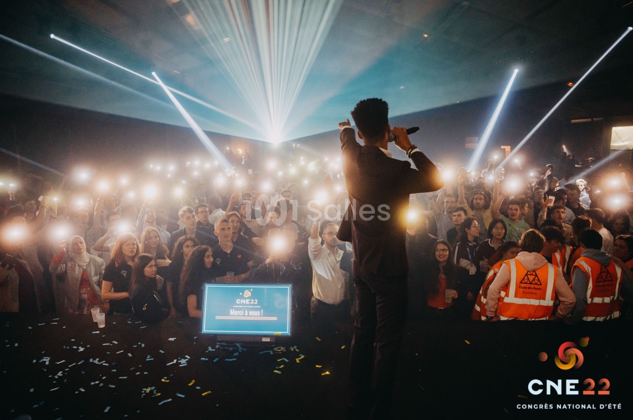 Un animateur sur scène devant une foule illuminée par des téléphones, lors d'un événement en intérieur.