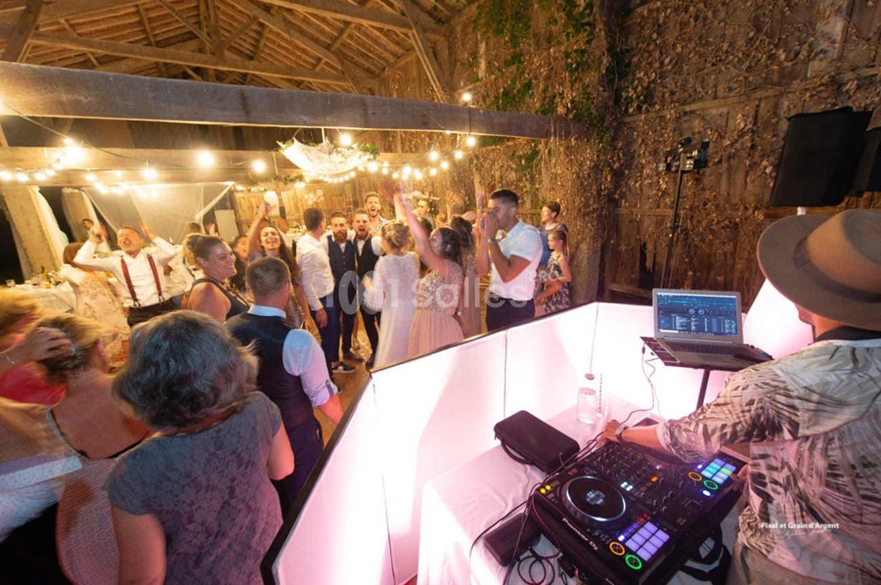 Groupe de personnes dansant sous des guirlandes lumineuses dans une grange, avec un DJ en arrière-plan.