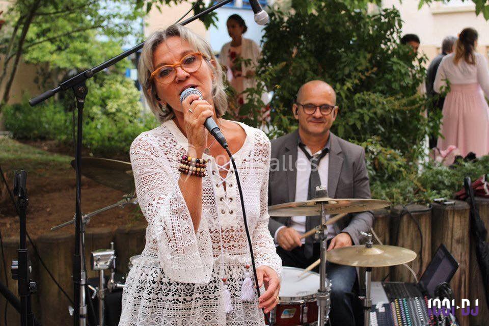 Une femme chante au micro devant un batteur, dans un cadre extérieur avec des arbres en arrière-plan.