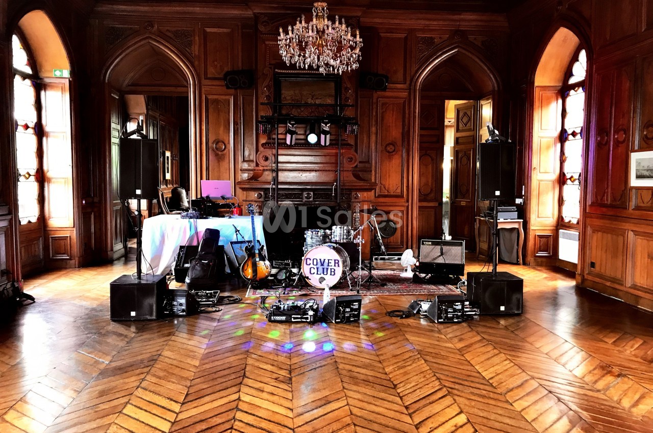 Équipement musical installé dans une salle en bois avec parquet, lustre et instruments prêts pour une performance.