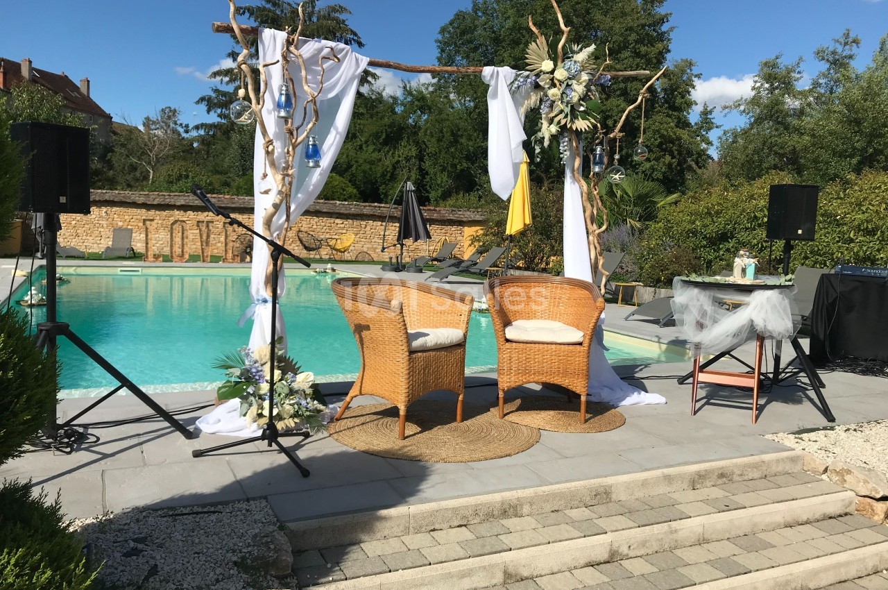 Deux fauteuils en rotin sous une arche décorée, installés près d'une piscine dans un cadre extérieur ensoleillé.