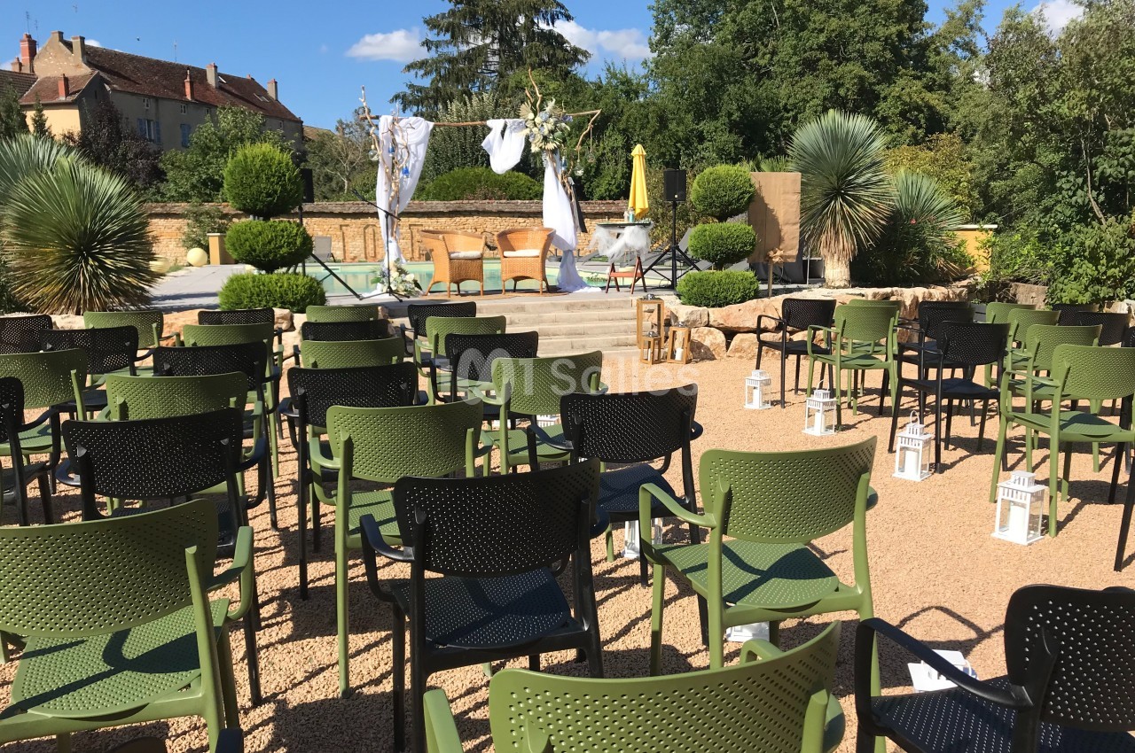 Chaises disposées en rangées face à une scène extérieure décorée, entourée de végétation et de bâtiments en arrière-plan.