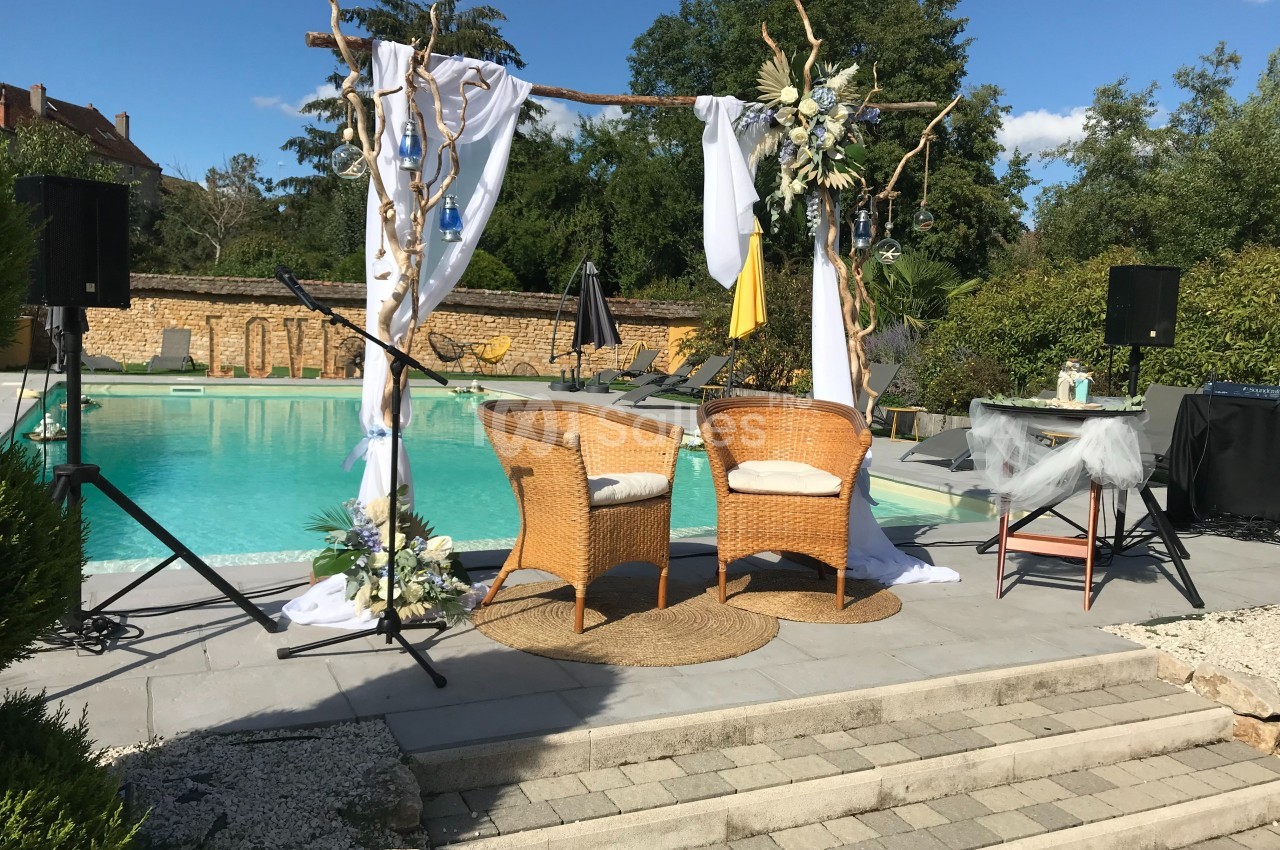 Deux fauteuils en rotin sous une arche décorée, installés près d'une piscine dans un cadre extérieur ensoleillé.