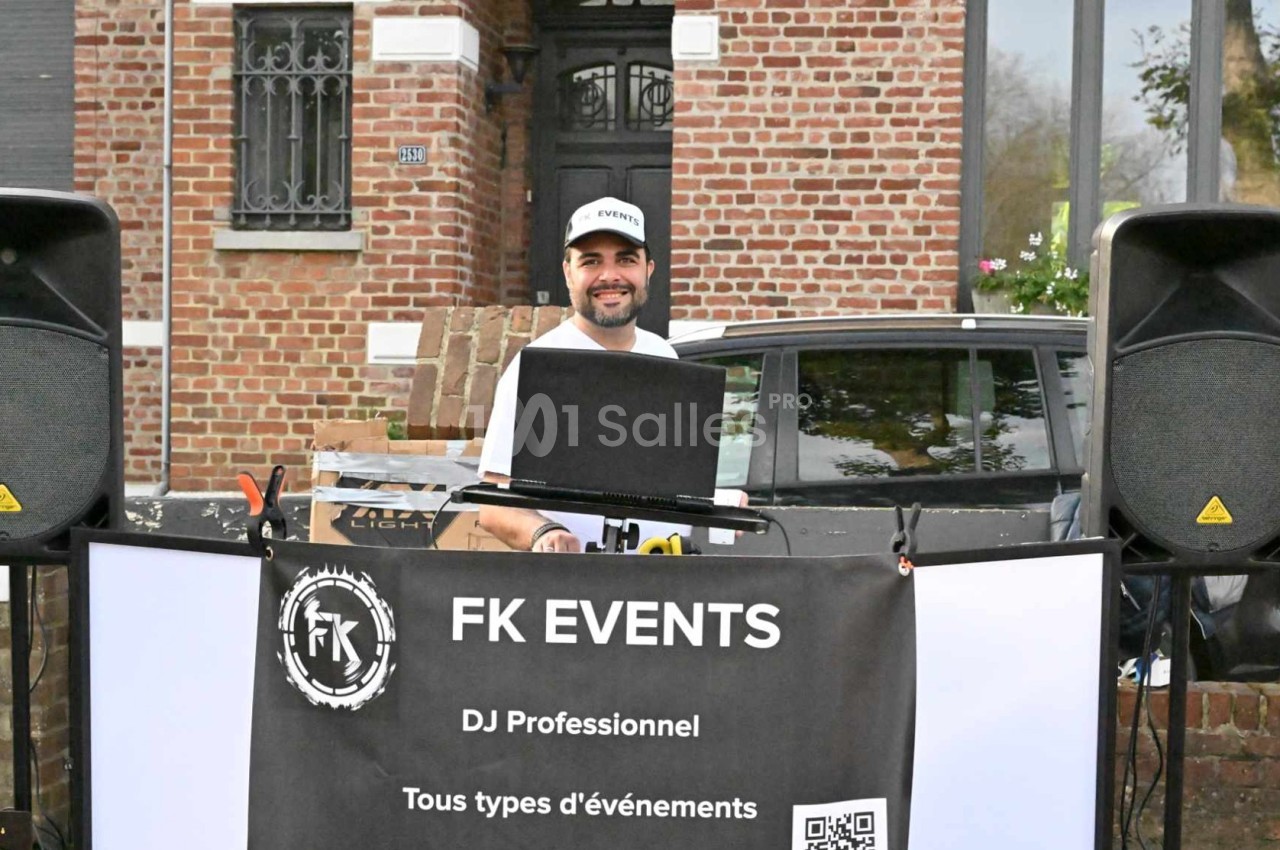 Un DJ souriant mixe en extérieur devant une maison en briques, entouré de matériel audio et d'une bannière promotionnelle.